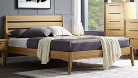 A wooden bed frame with dark bedding, pillows, and a light throw blanket, situated in a contemporary bedroom with large windows and natural light.